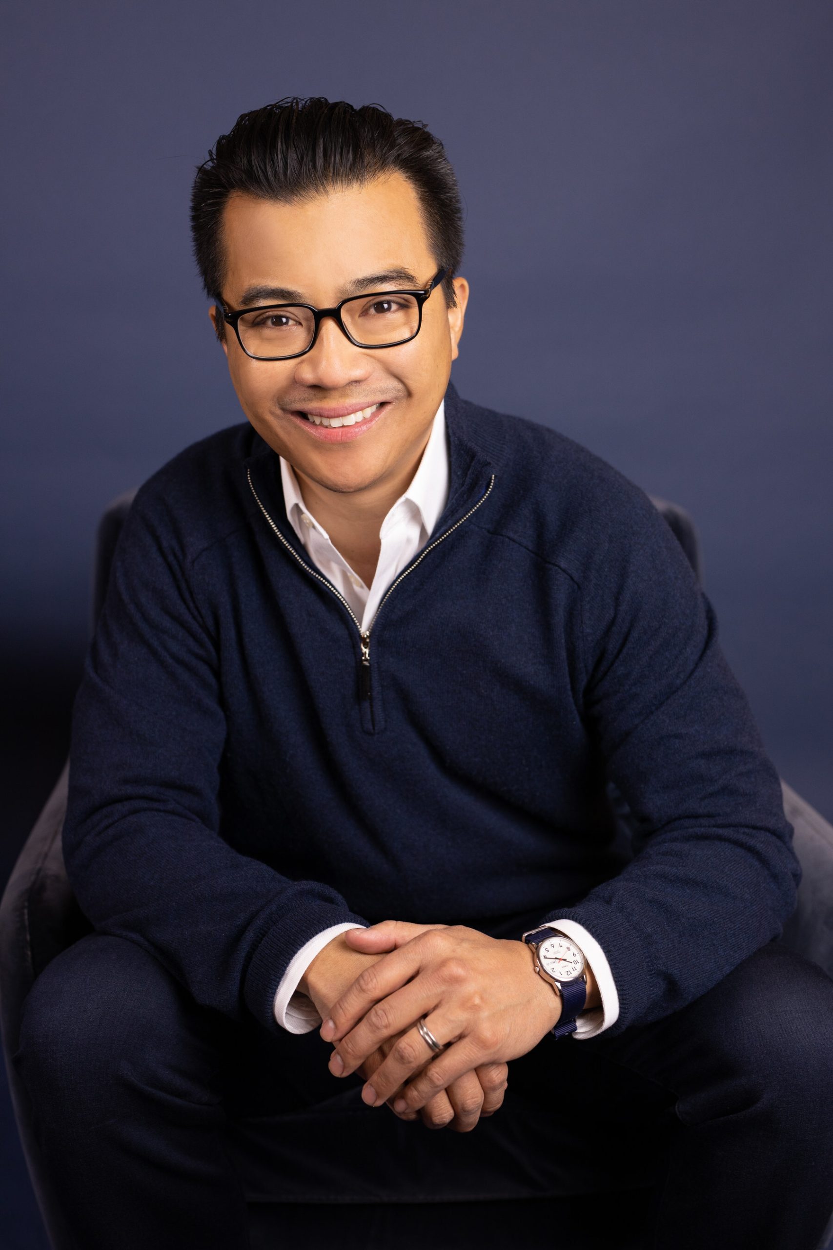 Smiling man with dark hair and glasses, wearing a navy blue half-zip sweater and white collared shirt, sitting with hands clasped.