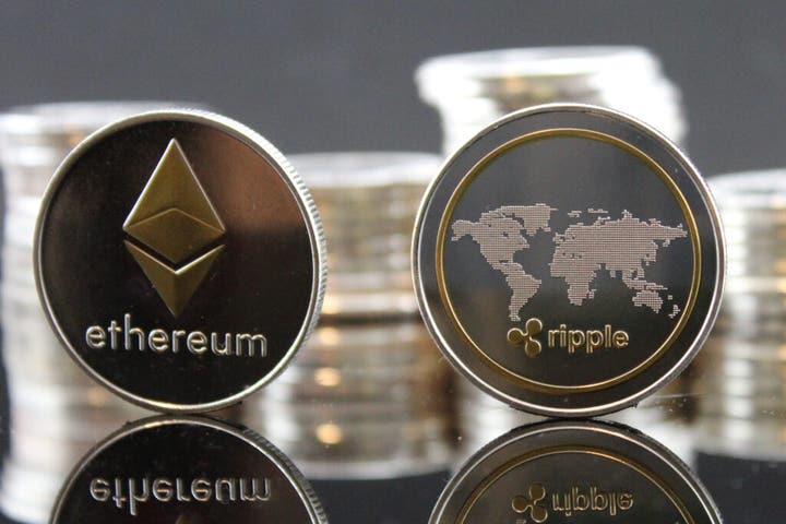 Ethereum and Ripple cryptocurrency coins displayed side by side on a reflective surface, with blurred stacks of coins in the background.